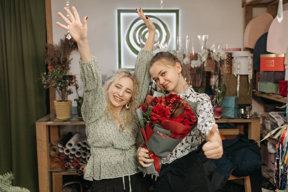 Two women in a floral shop, one holding a large bouquet of vibrant red roses wrapped in grey paper with red ribbon, both smiling and making hand gestures of celebration. The woman on the right has lig