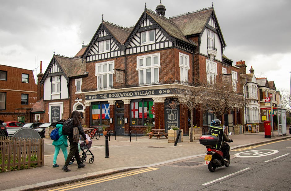 A traditional red-brick pub with black and white Tudor-style decorative timbering on the upper floors, situated on a busy street in East Sheen. The pub, named 'The Rookwood Village,' features a black 