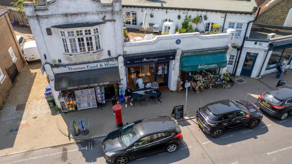 An aerial view of a charming street scene featuring a row of storefronts including Thames Food & Wine and La Petite. In front of the shops, a sidewalk displays a well-arranged bouquet of fresh flowers