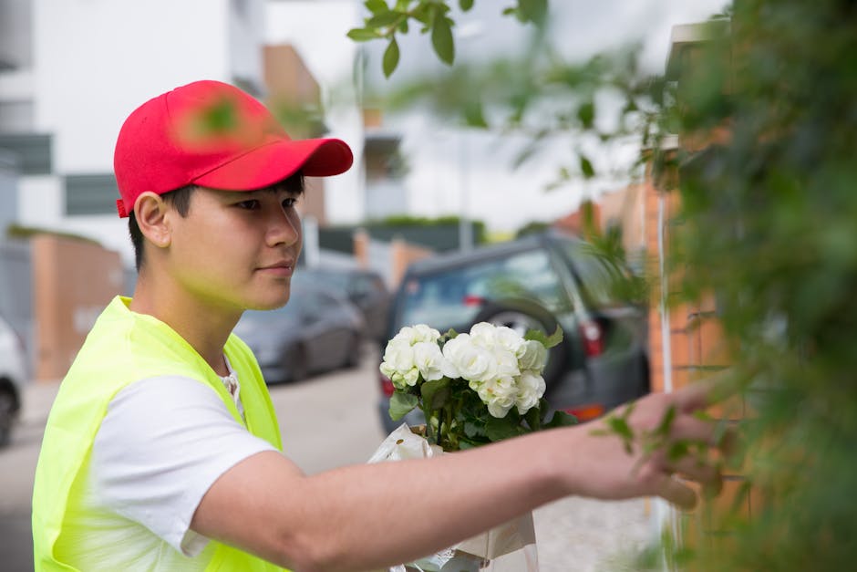 A young male florist wearing a red cap and a neon yellow vest, holding a bouquet of white hydrangeas wrapped in white paper and presenting it against a street background with parked cars and residenti