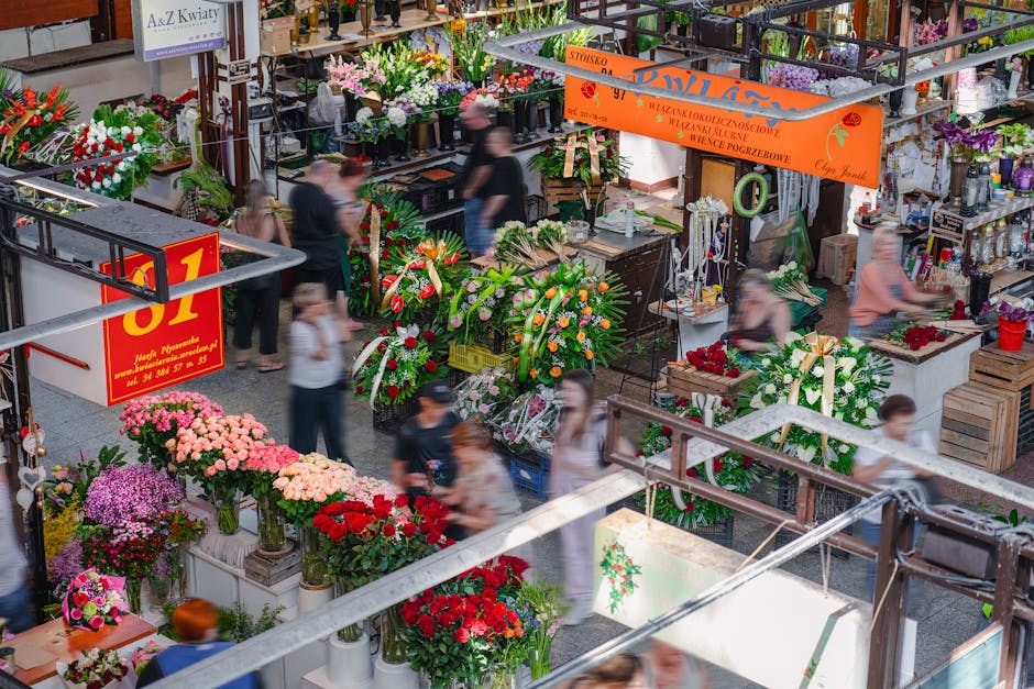 A vibrant indoor flower market featuring a variety of floral arrangements and bouquets displayed on tables and shelves. The flowers include pink, red, purple, and white chrysanthemums, roses, and lili